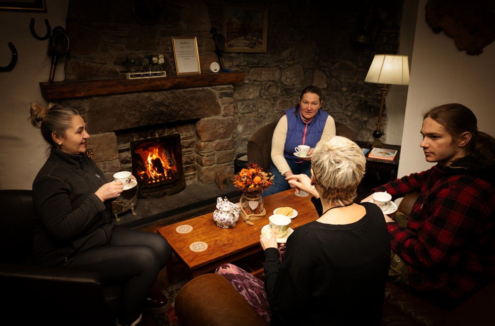 A group of people relaxing with tea and biscuits by the stone fireplace at The Old Inn, providing a warm welcome for NC500 travellers and hikers.
