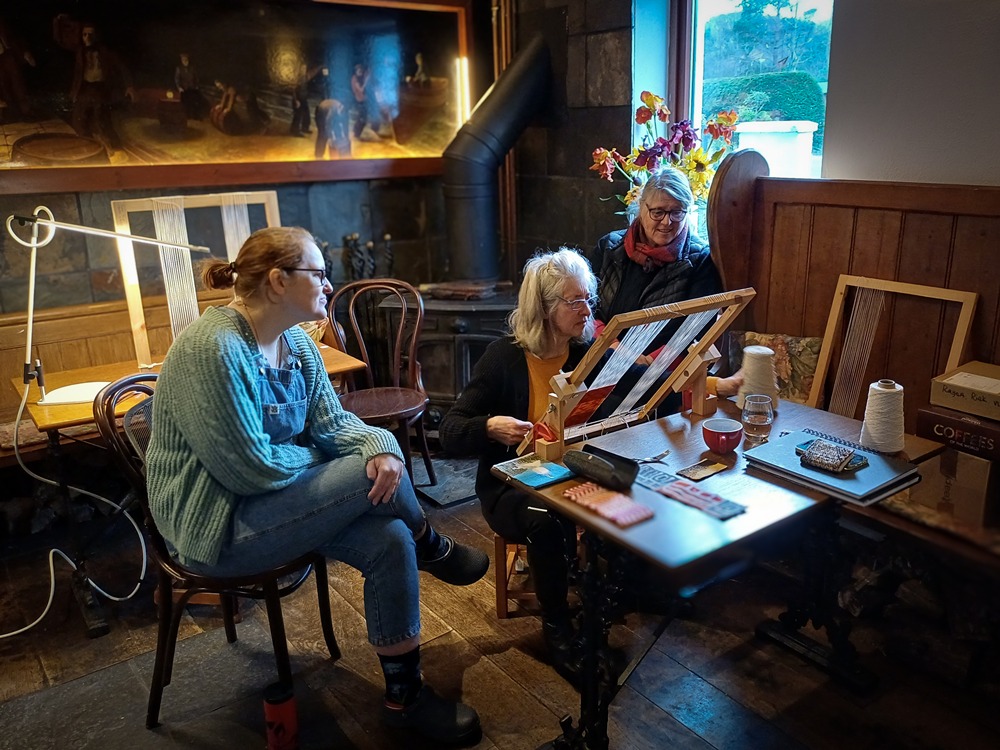A group of women attending a tapestry weaving workshop in a cosy, rustic studio.