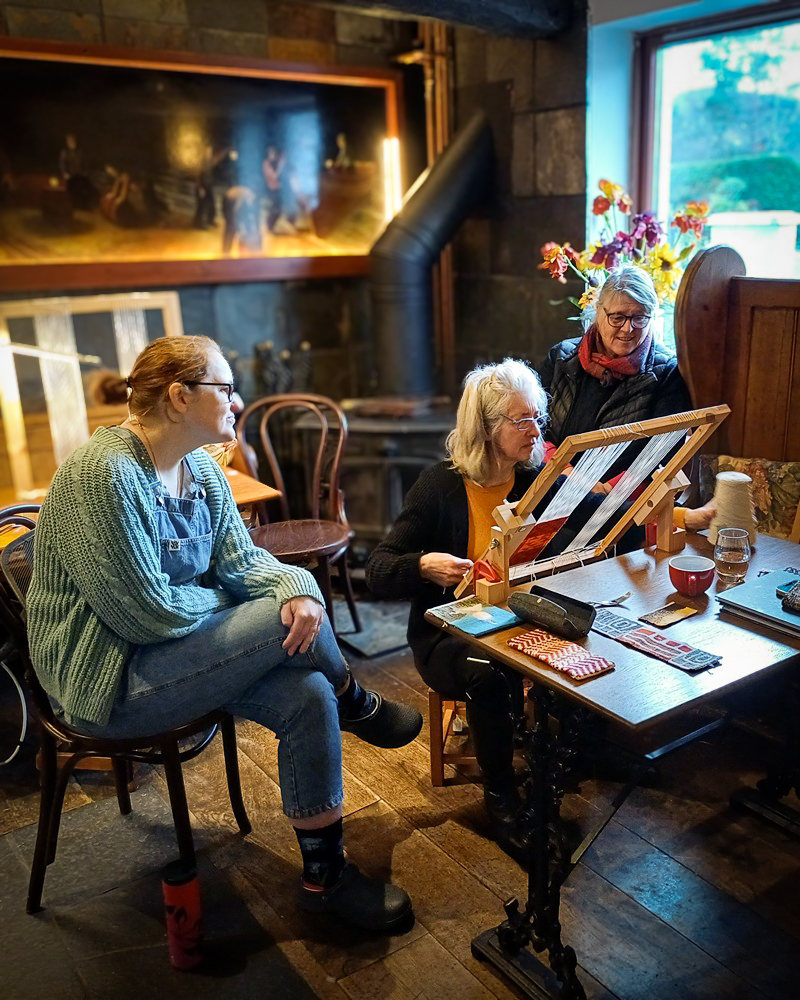 Three women at a tapestry weaving workshop in a rustic room. One works on a small tabletop loom while the others observe and chat. The background features a wood-burning stove and a large painting of a traditional workshop.