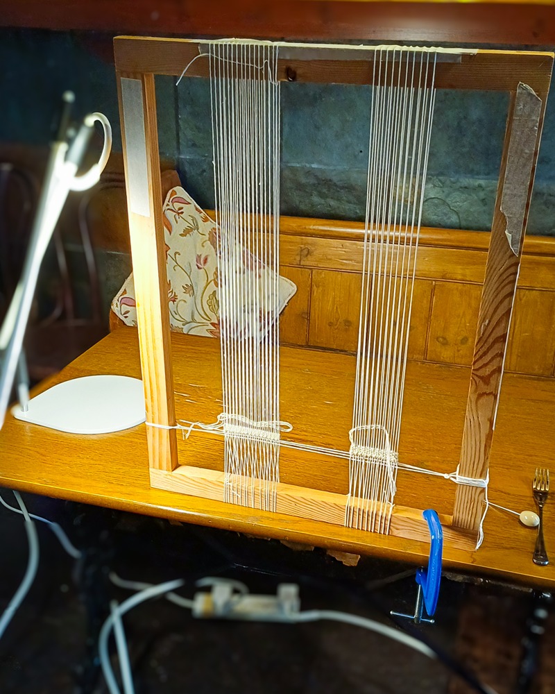 A close-up of a wooden tabletop frame loom clamped to a wooden desk. White warp threads are strung vertically across the frame, with the beginnings of a woven tapestry visible at the bottom.