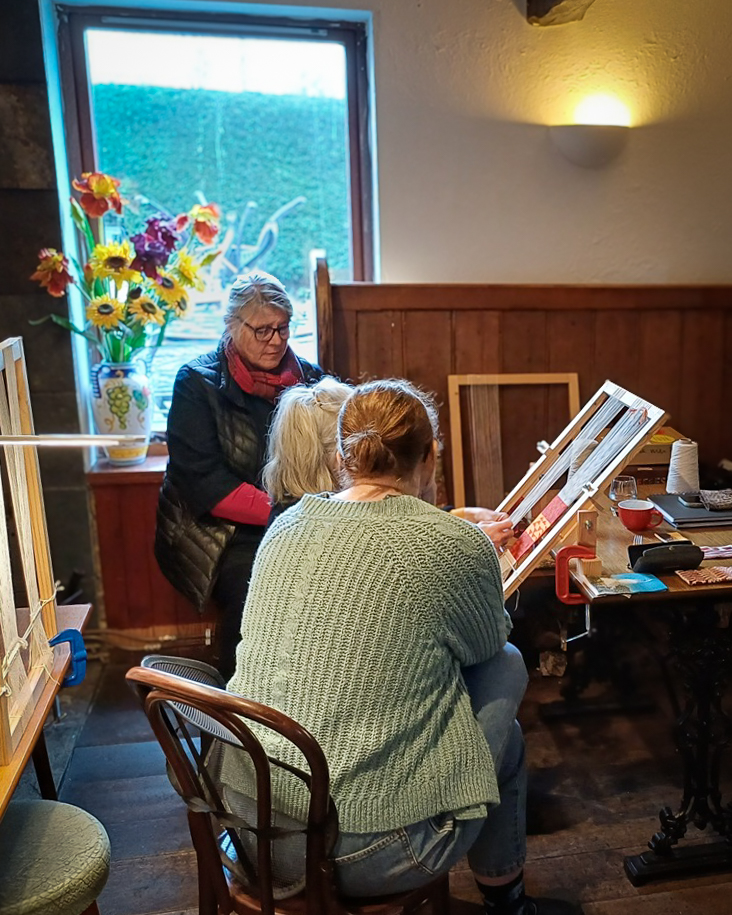 A rear view of three women gathered around a wooden table during a weaving workshop. One woman, wearing a light-green cable-knit jumper, sits in the foreground watching another participant work on a tilted wooden loom strung with red and white yarn.