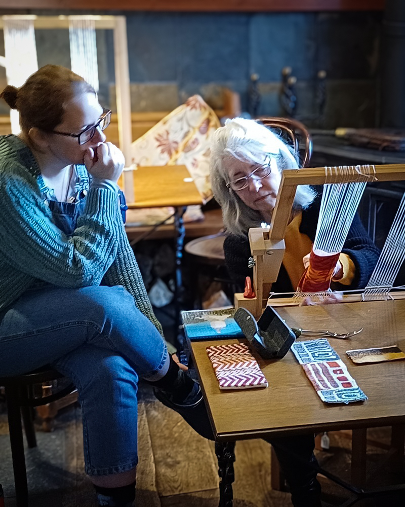 Two women concentrating on a weaving project at a wooden table. One operates a small timber loom while the other observes the technique. Several woven textile samples lie on the table in the foreground.
