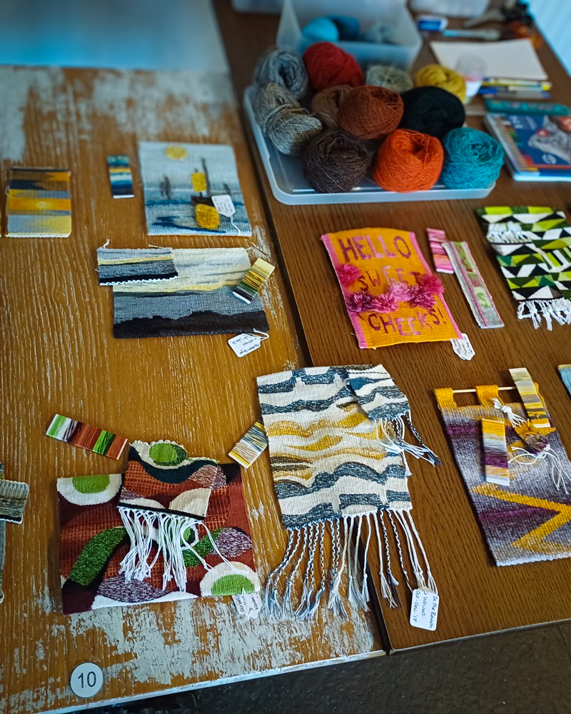 A collection of small, finished tapestry weavings and yarn samples laid out on a rustic wooden table, alongside several balls of multi-coloured wool.