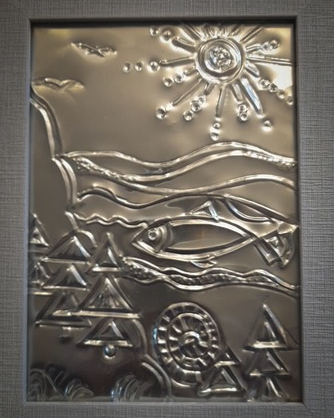 A rectangular piece of embossed pewter artwork in a light grey frame, depicting a stylised coastal landscape. The shimmering metal relief features a large sun with radiating rays, two birds in flight, a leaping fish in the water, and a foreground of triangular evergreen trees and a spiral shell.