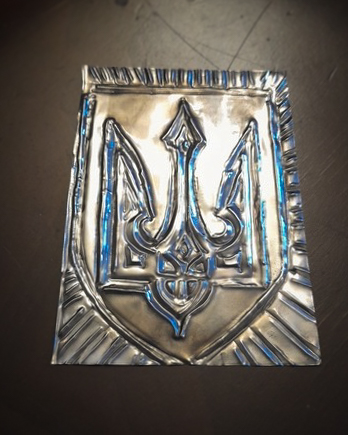 A close-up of a hand-crafted metal embossing on a silver-coloured sheet, showing the Tryzub, or coat of arms of Ukraine. The three-pronged trident symbol is raised in high relief against a hammered background, with visible tool marks and decorative crimped edges along the top and sides of the metal foil.