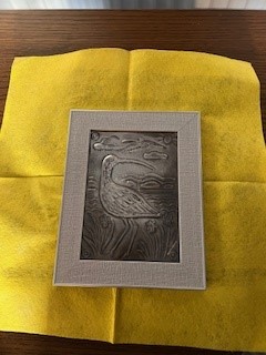A small, rectangular pewter embossing in a light grey frame, resting on a yellow cloth. The metal relief depicts a curlew with its characteristic long, curved beak standing in marshland with reeds and flowers. In the background, stylised hills and clouds are visible under a simple sky.