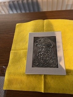 A small, framed pewter embossing of an eagle's head in profile. The intricate metal relief showcases highly detailed feathers and a sharp beak, resting on a bright yellow cloth over a wooden surface.