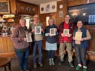Five smiling participants stand in a wood-panelled room, each holding up a small, framed piece of embossed metal artwork they created during a workshop. The group includes men and women of various ages, proudly displaying different designs including wildlife and symbols.