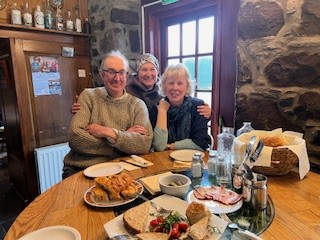 Three workshop participants sitting around a circular wooden table during a lunch break in a rustic room with stone walls. They are smiling at the camera, with a spread of sandwiches, salad, and bread rolls laid out before them.