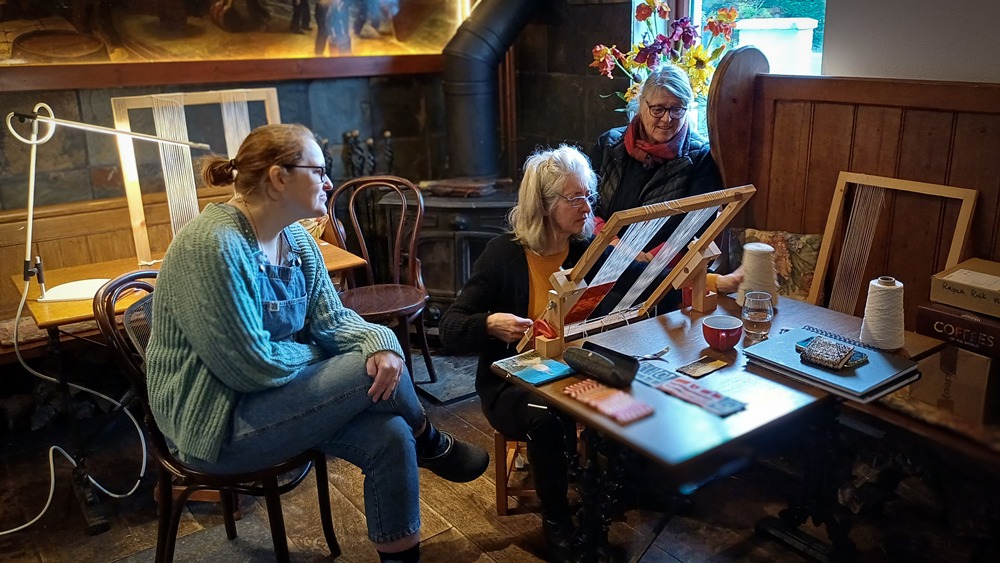Three women participate in a weaving workshop at a wooden table. The rustic room includes several looms, a wood-burning stove, and a vase of sunflowers by the window.
